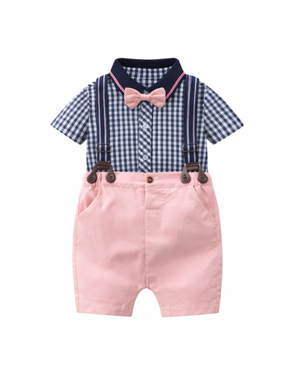 ネイビー×白のギンガムチェック半袖シャツに、ピンクの蝶ネクタイとネイビーのサスペンダー付きピンクショートパンツを組み合わせたベビー用セットアップが、白背景で正面から平置き撮影されている。
