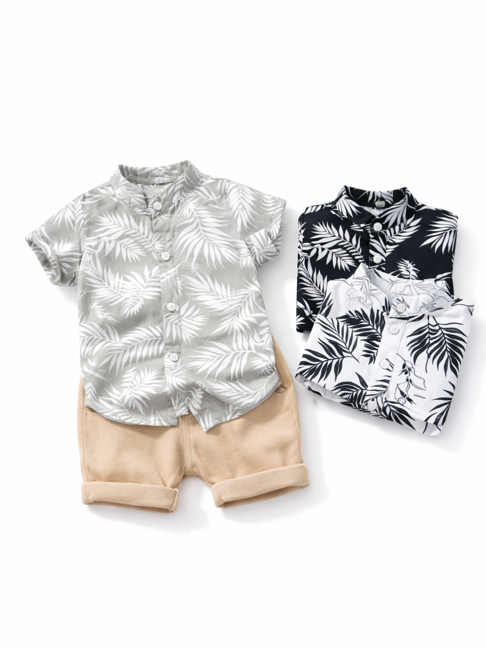 白背景のスタジオ撮影で、子ども用の夏服セット（グレーのボタンダウン半袖シャツ＋ベージュのショートパンツ）をフラットレイで配置し、右側にモノトーンのリーフ柄シャツ2枚を折りたたんで並べた商品写真。
