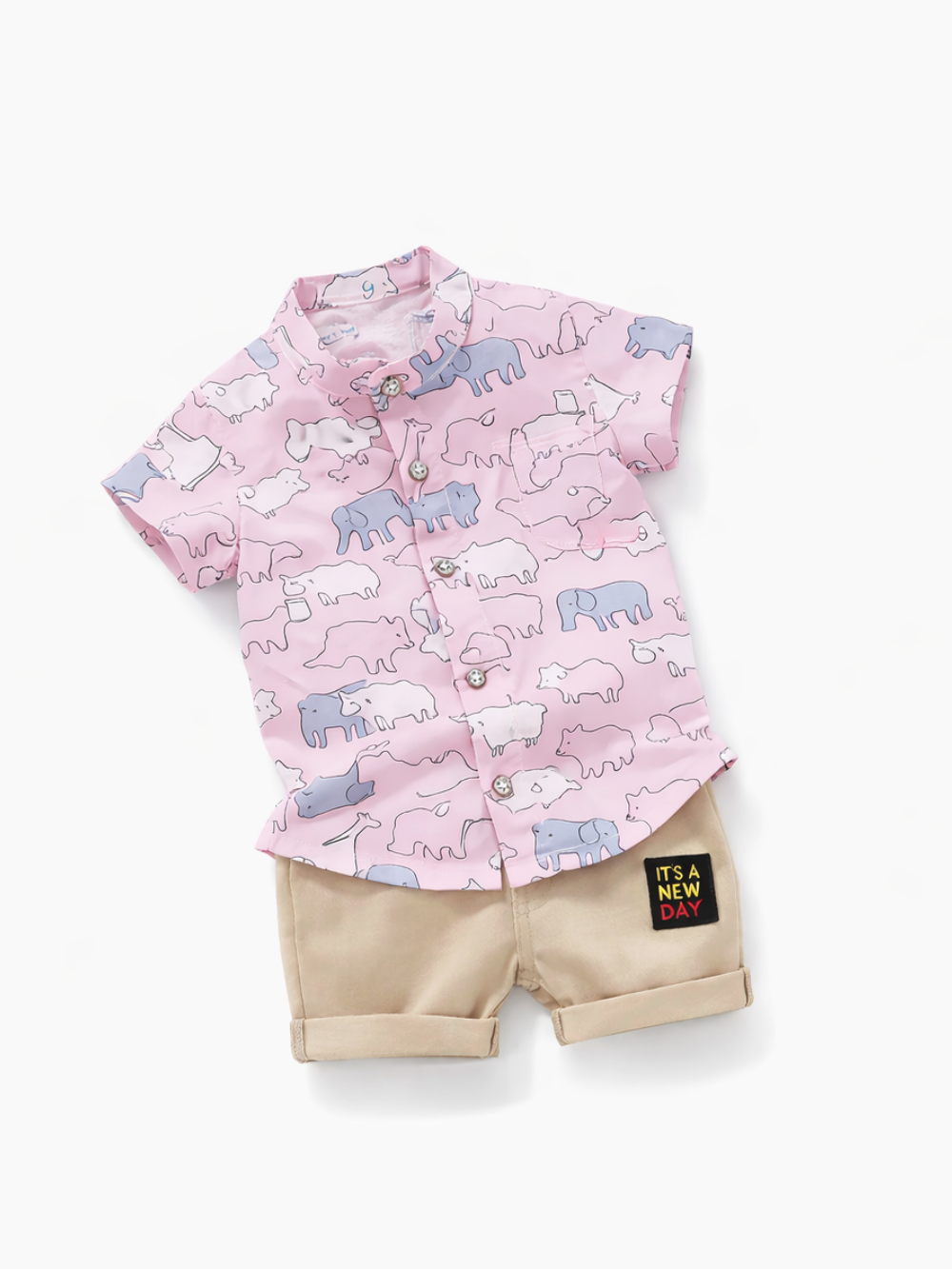 白背景に平置きされた子ども用上下セット。動物柄のピンク半袖ボタンシャツと、ベージュのショートパンツ（「IT’S A NEW DAY」パッチ付き）。
