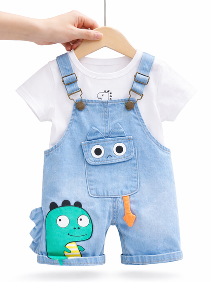白背景に、子ども用の上下セットがハンガーに掛けられている。白い半袖Tシャツの上に、ライトブルーのデニムサロペット（ショート丈）を重ねたデザイン。サロペットの胸ポケットには目の付いたキャラクター風のアップリケがあり、左脚にはグリーンの恐竜イラスト、中央にはオレンジ色の小さな飾りが付いている。木製ハンガーを手で持って吊るした状態で、全体が明るく均一な光で撮影されている。```
