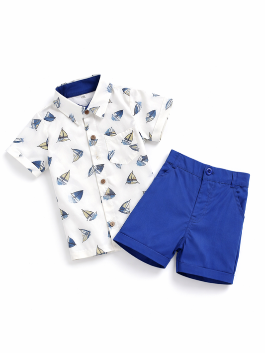 白背景に、子ども用の上下2点セットが平置きで配置されている。上は白地の半袖ボタンシャツで、青とベージュのヨット柄が全体にプリントされ、木目調ボタンと胸ポケットが付いている。下はロイヤルブルーのショートパンツで、前ボタンとベルトループがあり、裾は折り返し仕上げ。全体が明るく均一な光で撮影され、輪郭がはっきり見える。```
