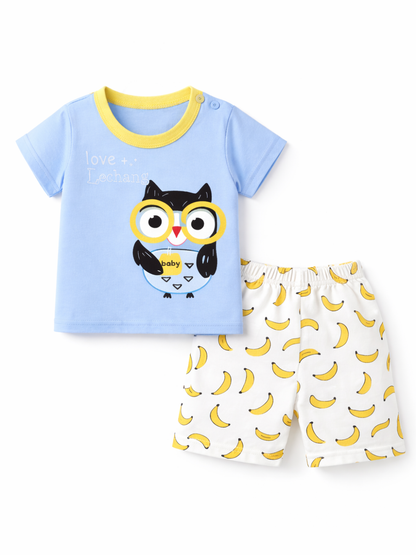 白背景のスタジオ撮影で、子ども用上下セットをフラットレイで配置。イエローの襟がアクセントのライトブルーTシャツにフクロウプリント、ホワイトのバナナ柄ショートパンツを組み合わせたデザイン。
