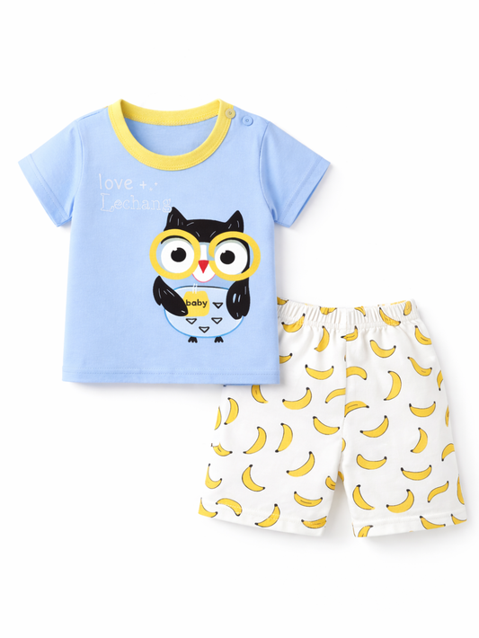 白背景のスタジオ撮影で、子ども用上下セットをフラットレイで配置。イエローの襟がアクセントのライトブルーTシャツにフクロウプリント、ホワイトのバナナ柄ショートパンツを組み合わせたデザイン。
