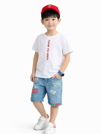 白背景のスタジオ撮影で、赤いキャップをかぶった男の子が白いプリントTシャツとパッチ付きデニムハーフパンツを着用し、白いスニーカーで立ち姿のポーズをしている。
