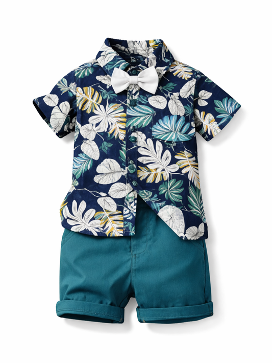 白背景のスタジオ風商品写真。男の子用の上下セットが正面向きに配置されている。ネイビー地に白・グリーン・イエロー系のボタニカル柄が入った半袖シャツに、首元に白い蝶ネクタイが付いている。下は深いティールカラーのショートパンツで、裾を折り返したデザイン。
