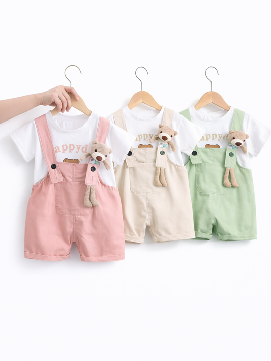 白背景のスタジオ風商品写真。ベビー用のサロペットセットが3着、木製ハンガーに掛けて並んでいる。白い半袖Tシャツに、ピンク・ベージュ・ミントグリーンのサロペットショーツを組み合わせたデザインで、胸元に小さなポケット付き。各サロペットの肩ひもには、ストライプのマフラーを巻いたぬいぐるみのクマチャームが付いている。左端のハンガーは手で持たれている。
