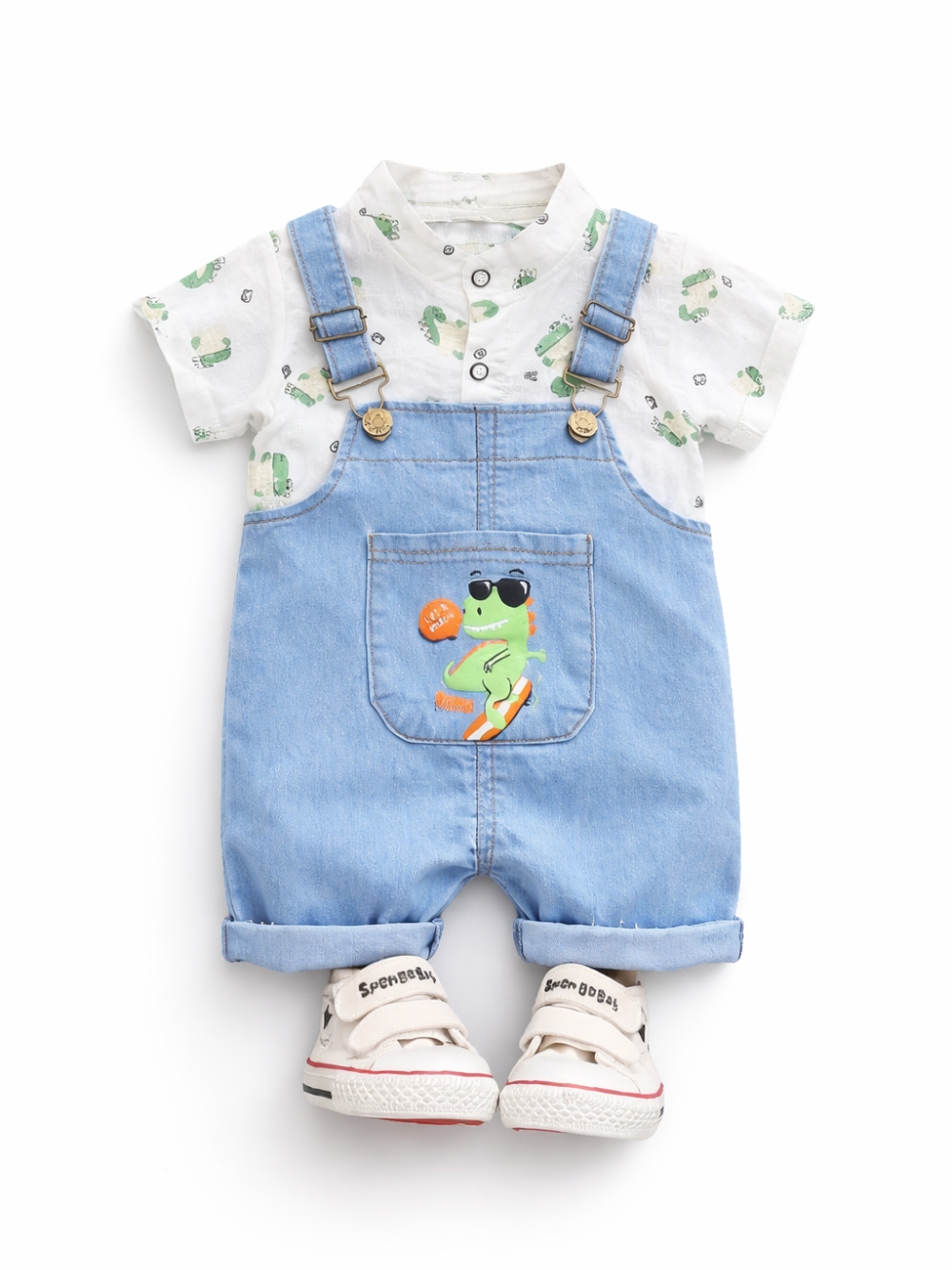 白背景のスタジオ風商品写真。ベビー用コーデが正面向きに配置されている。白い半袖シャツは小さな恐竜柄の総柄プリントで、前立てボタン付き。上からライトブルーのデニムサロペットを重ね、肩ひもは金具と調節バックル付き。胸ポケットにはサングラスをかけた恐竜のイラストが入っている。足元に白い面ファスナー付きスニーカーが一足置かれている。

