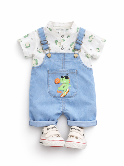 白背景のスタジオ風商品写真。ベビー用コーデが正面向きに配置されている。白い半袖シャツは小さな恐竜柄の総柄プリントで、前立てボタン付き。上からライトブルーのデニムサロペットを重ね、肩ひもは金具と調節バックル付き。胸ポケットにはサングラスをかけた恐竜のイラストが入っている。足元に白い面ファスナー付きスニーカーが一足置かれている。
