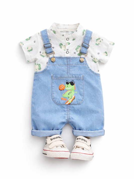 白背景のスタジオ風商品写真。ベビー用コーデが正面向きに配置されている。白い半袖シャツは小さな恐竜柄の総柄プリントで、前立てボタン付き。上からライトブルーのデニムサロペットを重ね、肩ひもは金具と調節バックル付き。胸ポケットにはサングラスをかけた恐竜のイラストが入っている。足元に白い面ファスナー付きスニーカーが一足置かれている。
