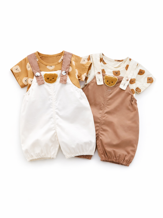 白背景のスタジオ風商品写真。ベビー用のロンパース風セットが2着、並べて配置されている。左はキャメル色のくま柄半袖トップスに、白のサロペットロンパースを重ねたデザインで、胸元にくま顔のワッペン付き。右はクリーム色のくま柄半袖トップスに、ブラウンのサロペットロンパースを重ねたデザインで、同じく胸元にくま顔ワッペンが付いている。靴などの小物は写っていない。
