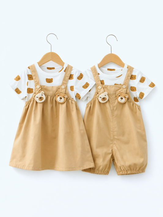白背景のスタジオ風商品写真。ベビー用の2コーデが木製ハンガーに掛けて並んでいる。どちらも白地にくま顔の総柄プリントの半袖Tシャツと、キャメル色のサロペットを組み合わせている。左はギャザー入りのジャンパースカート風デザイン、右はロンパース風のショート丈デザインで、胸元にくま顔の立体チャームが2つ付いている。
