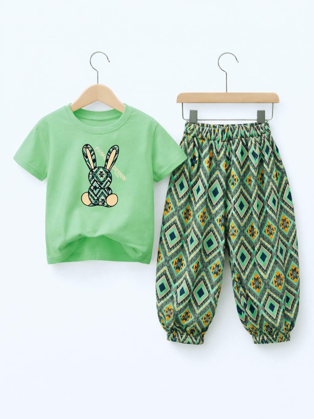 白背景のスタジオ風商品写真。子ども用の上下セットがハンガーに掛けて並べて表示されている。左にライトグリーンの半袖Tシャツが木製ハンガーに掛かり、中央に幾何学柄のウサギのイラストとカーブした文字デザインがプリント。右に同じ木製クリップハンガーで、グリーン系の幾何学模様（ダイヤ柄）のゆったりしたロングパンツが掛けられている。パンツはウエストと裾がゴム仕様。
