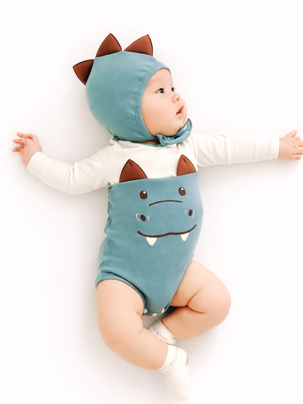 白背景のスタジオ風商品写真。ミントグリーンの恐竜モチーフのロンパースを着た赤ちゃんが、仰向けで腕を広げたポーズで寝ている。フードには茶色のトゲ状パーツが付き、ロンパース前面には恐竜の顔プリント（目と牙）がある。中にはオフホワイトの長袖インナーを着用し、白い靴下を履いている。
