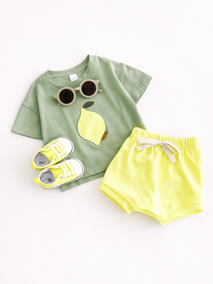 白背景のスタジオ風商品写真。子ども用の上下セットと小物がフラットに配置されている。セージグリーンの半袖Tシャツには、レモンのアップリケが付いており、首元に丸いサングラスが置かれている。左下に黄色のスニーカーが一足、右側に淡いイエローのショートパンツがあり、ウエストはゴム仕様で白いリボン付き。
