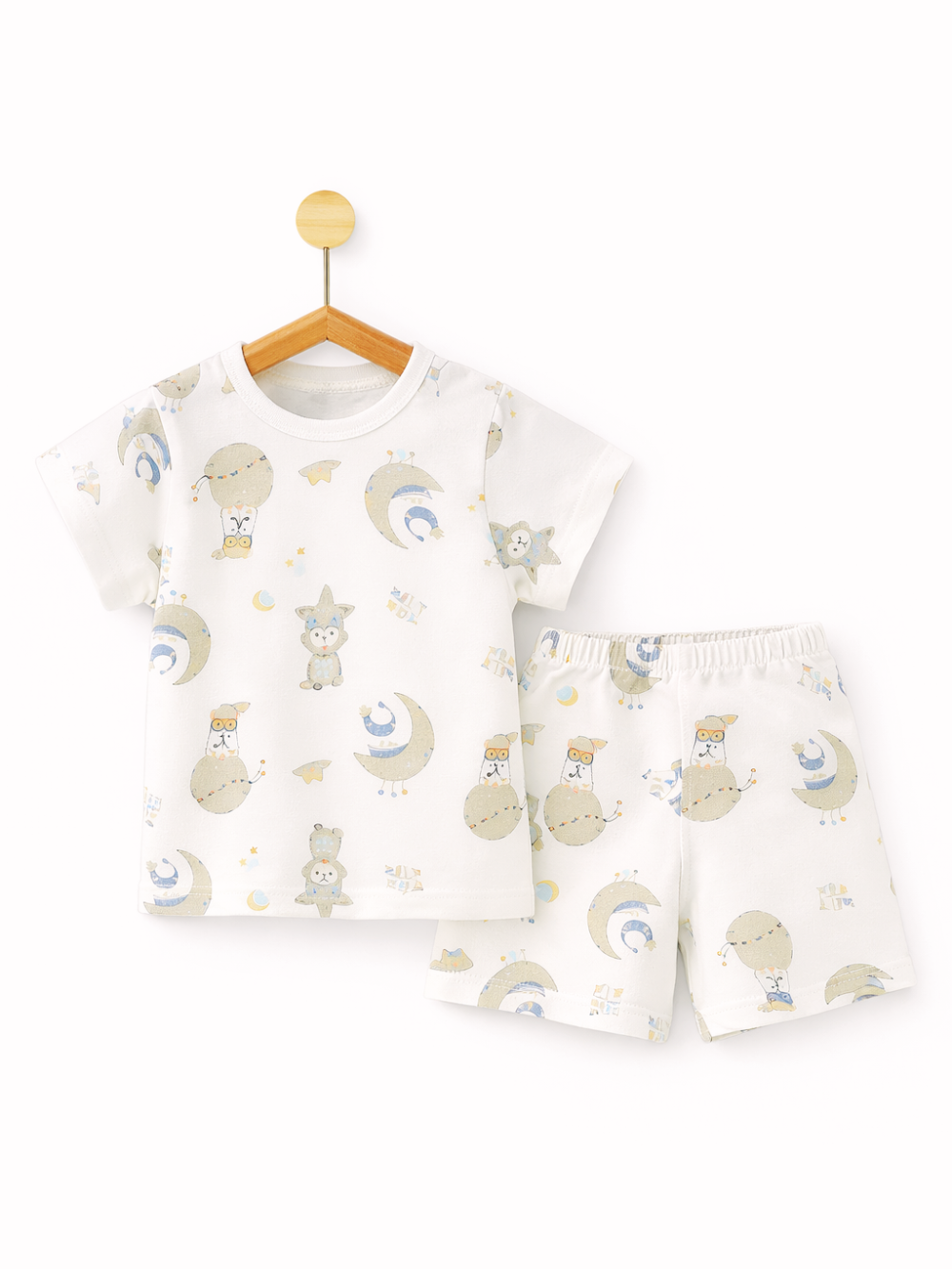 白背景のスタジオ風商品写真。子ども用の上下セットが配置されている。左に木製ハンガーに掛かった白い半袖Tシャツがあり、月や星、動物などの淡いパステル調イラストが総柄でプリントされている。右に同柄の白いショートパンツが置かれ、ウエストはゴム仕様。全体が明るくクリーンに撮影されている。
