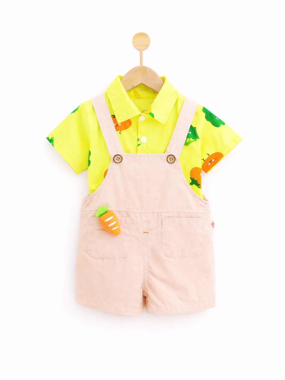 白い背景に木製ハンガーで吊るされた子ども用の上下セット。黄色の半袖シャツにはにんじんと木のイラストが散りばめられ、淡いピンクのサロペットショーツには前ポケットとにんじん型の飾りが付いている。靴などの小物は写っていない。
