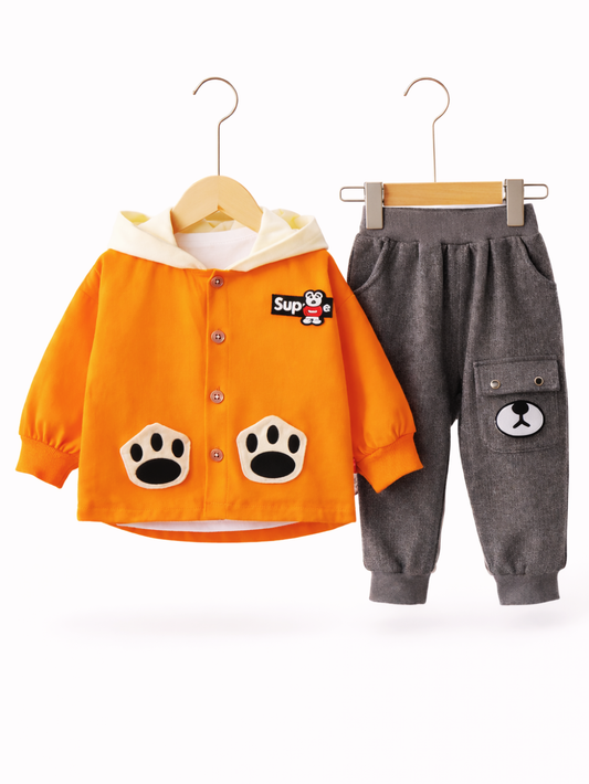 白い無地の背景に、子ども用の服が2点並んで掛けられている。左は木製ハンガーに掛かったオレンジ色のフード付きジャケットで、前ボタンと肉球モチーフのポケット、胸元に小さなワッペンがある。右はクリップ付きハンガーに掛かったダークグレーのジョガーパンツで、サイドポケットのフラップにクマの顔のパッチが付いている。
