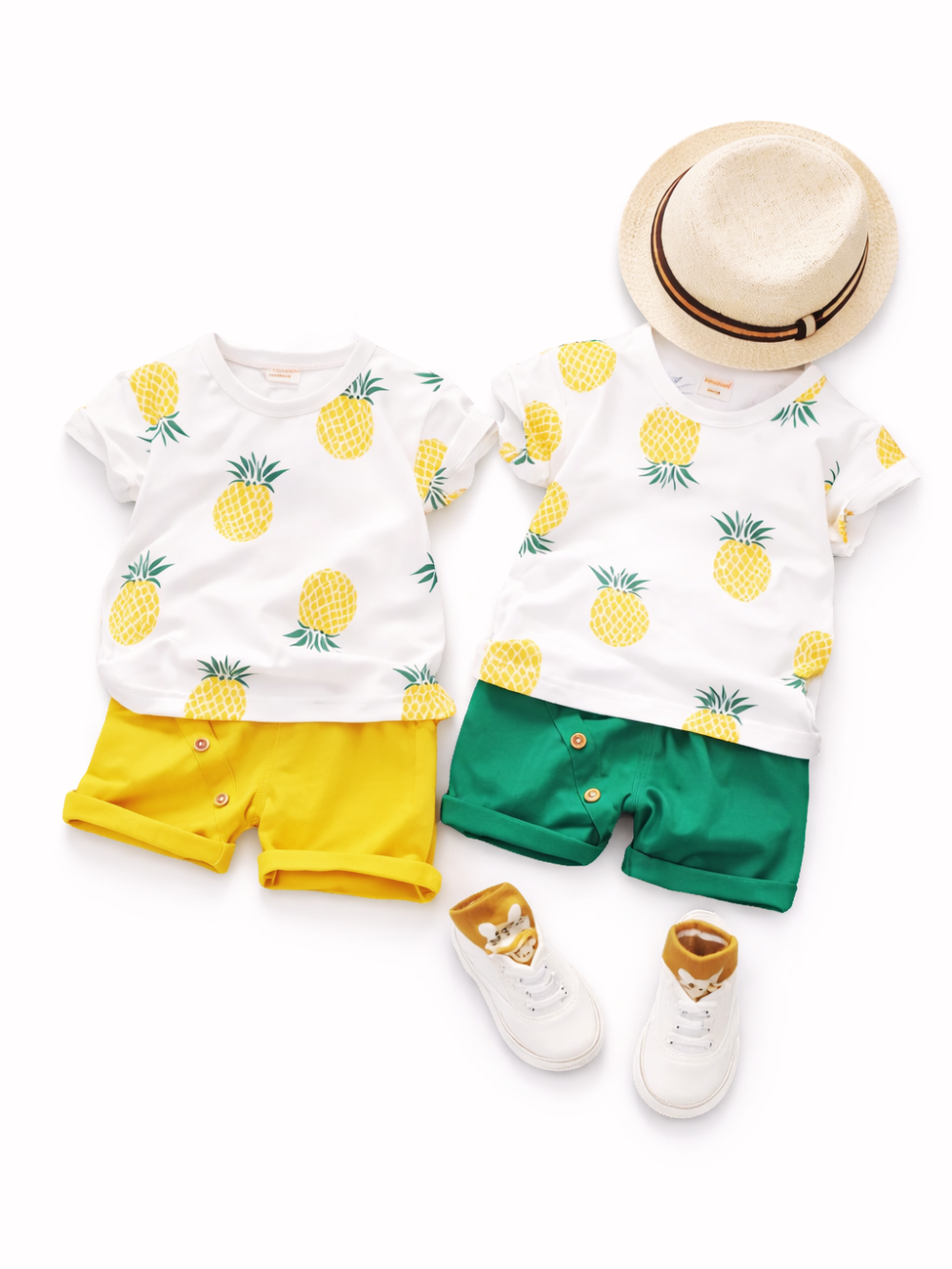 白背景に並べられた子ども用の夏服2セット。パイナップル柄の白Tシャツに、左は黄色のショートパンツ、右は緑のショートパンツ。右上に麦わら帽子、下部に白いスニーカーと茶色のソックスが置かれている。
