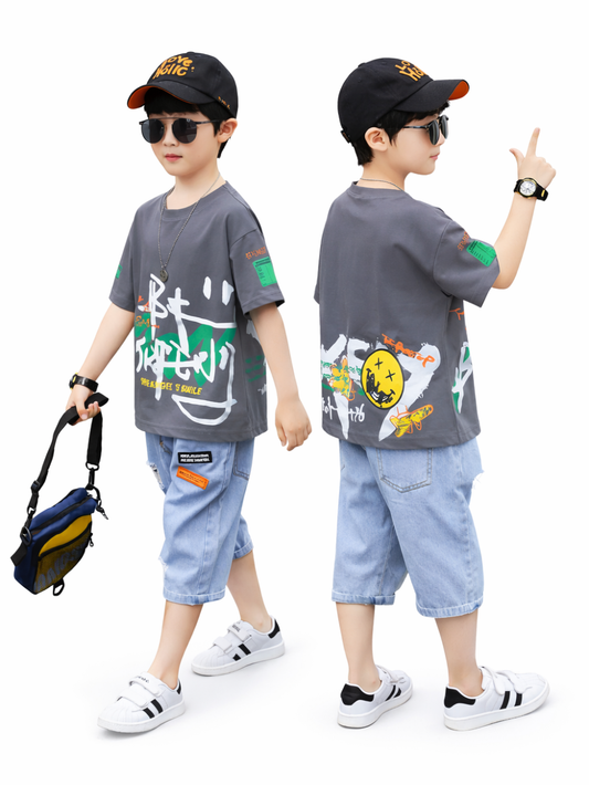 白背景のスタジオ撮影。グレーのグラフィックTシャツとライトブルーのデニムハーフパンツを着た男の子が、正面寄りと背面寄りの2ポーズで並んでいる。黒いキャップとサングラス、腕時計を着用し、片手に小さなショルダーバッグを持っている。足元は白いスニーカー。
