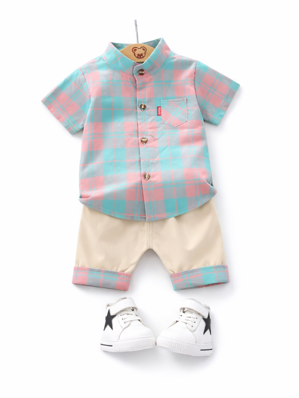 白背景のスタジオ撮影。淡いピンク×ミントのチェック柄の半袖ボタンシャツと、裾に同柄カフスが付いたベージュのショートパンツをハンガーに整えて配置し、下に黒い星アクセントの白いハイカットスニーカーを並べたフラットレイ画像。

