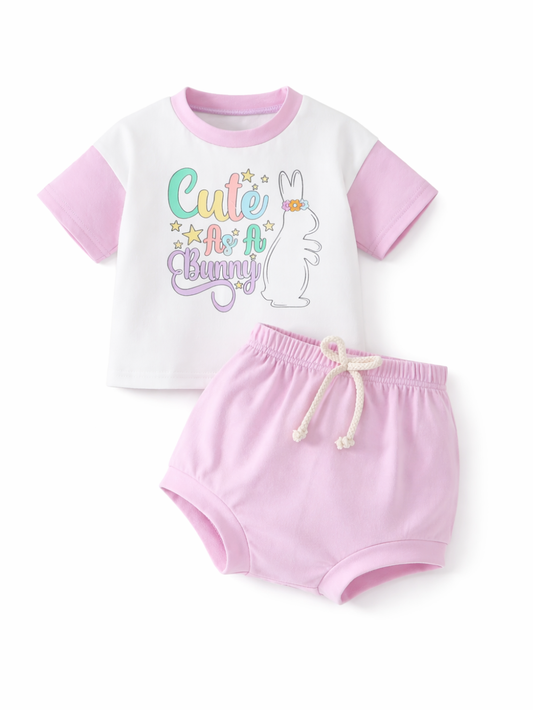 白い背景の上に、ベビー用のツーピースコーデがフラットに配置されている。白地に淡いピンクの切り替えが入った半袖Tシャツには「Cute As A Bunny」の文字とウサギのイラストがあり、下には淡いピンクのブルマ風ショーツが並ぶ。ショーツはウエストゴムで、生成りのひもが付いている。
