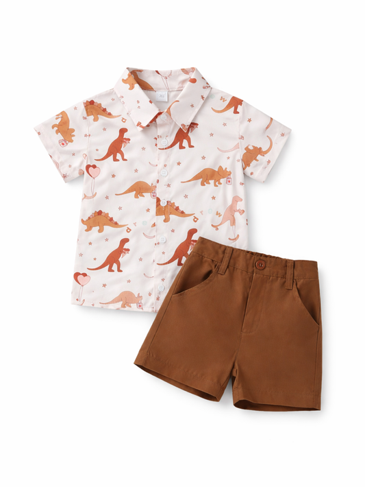 白背景の上に、子ども用の上下2点セット（恐竜柄の半袖ボタンシャツとブラウンのショートパンツ）が平置きで整えて配置されている。シャツは白地に茶系の恐竜と小さな星柄が散り、パンツはシンプルな無地で、自然な陰影が入ったカタログ風の商品写真。
