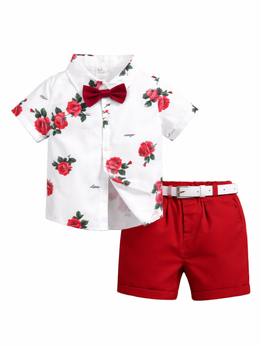 白背景に、子ども用の上下2点セットが平置きで配置されている。上は白地の半袖ボタンシャツで、赤いバラと緑の葉の花柄プリントが全体に入っており、胸元にワインレッドの蝶ネクタイが付いている。下は鮮やかな赤のショートパンツで、白いベルトとシルバーのバックルが付属。全体が均一な光で撮影され、輪郭がはっきり見える。
