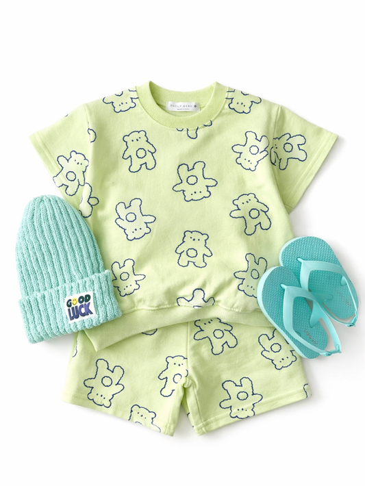 白背景に、子ども用の上下セットが平置きで配置されている。淡いライムグリーンの半袖トップスとショートパンツで、全体にネイビーのくま風イラストが散りばめられている。左側にミントカラーのニット帽が置かれ、「GOOD LUCK」のワッペンが付いている。右側にはミント色のビーチサンダルが添えられている。全体が均一な光で撮影され、輪郭がはっきり見える。
