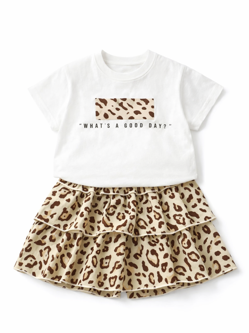 白背景に、子ども用の上下2点セットが平置きで配置されている。上は白い半袖Tシャツで、胸元にヒョウ柄の四角いパッチと「WHAT’S A GOOD DAY?」の黒い文字プリントが入っている。下はベージュ×ブラウンのヒョウ柄ティアードスカートで、フリルが重なった段々デザイン。全体が均一な光で撮影され、素材の質感と輪郭がはっきり見える。
