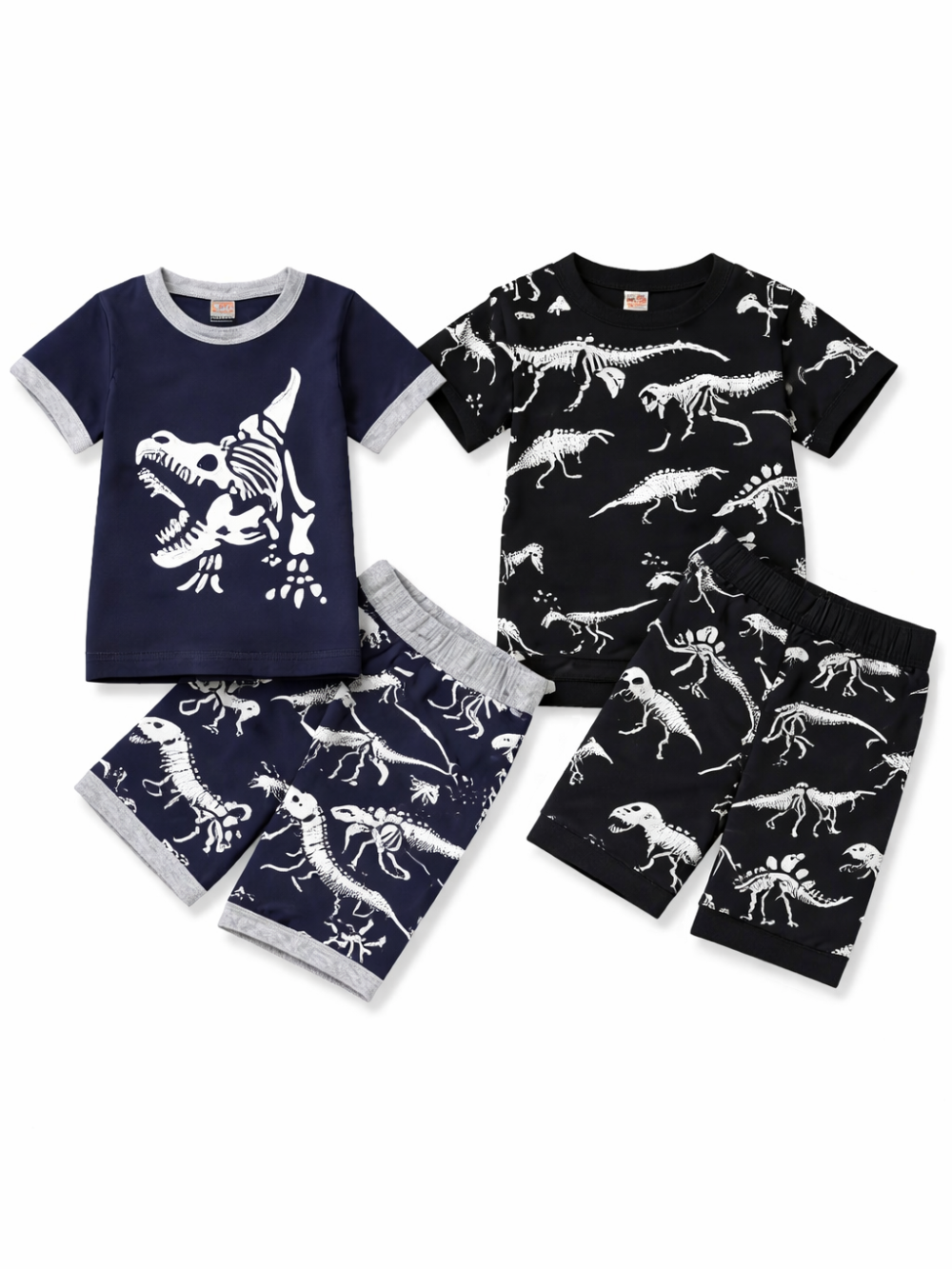 白背景に、子ども用の恐竜柄上下2点セットが2種類並べて平置きで配置されている。左はネイビーの半袖Tシャツに白いティラノサウルス骨格プリント、同柄のネイビーショートパンツがセット。右は黒の半袖Tシャツに白い恐竜骨格の総柄プリント、同柄の黒いショートパンツがセット。全体が均一な光で撮影され、柄と輪郭がはっきり見える。
