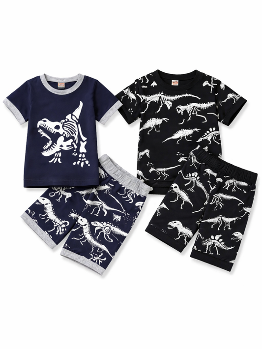 白背景に、子ども用の恐竜柄上下2点セットが2種類並べて平置きで配置されている。左はネイビーの半袖Tシャツに白いティラノサウルス骨格プリント、同柄のネイビーショートパンツがセット。右は黒の半袖Tシャツに白い恐竜骨格の総柄プリント、同柄の黒いショートパンツがセット。全体が均一な光で撮影され、柄と輪郭がはっきり見える。
