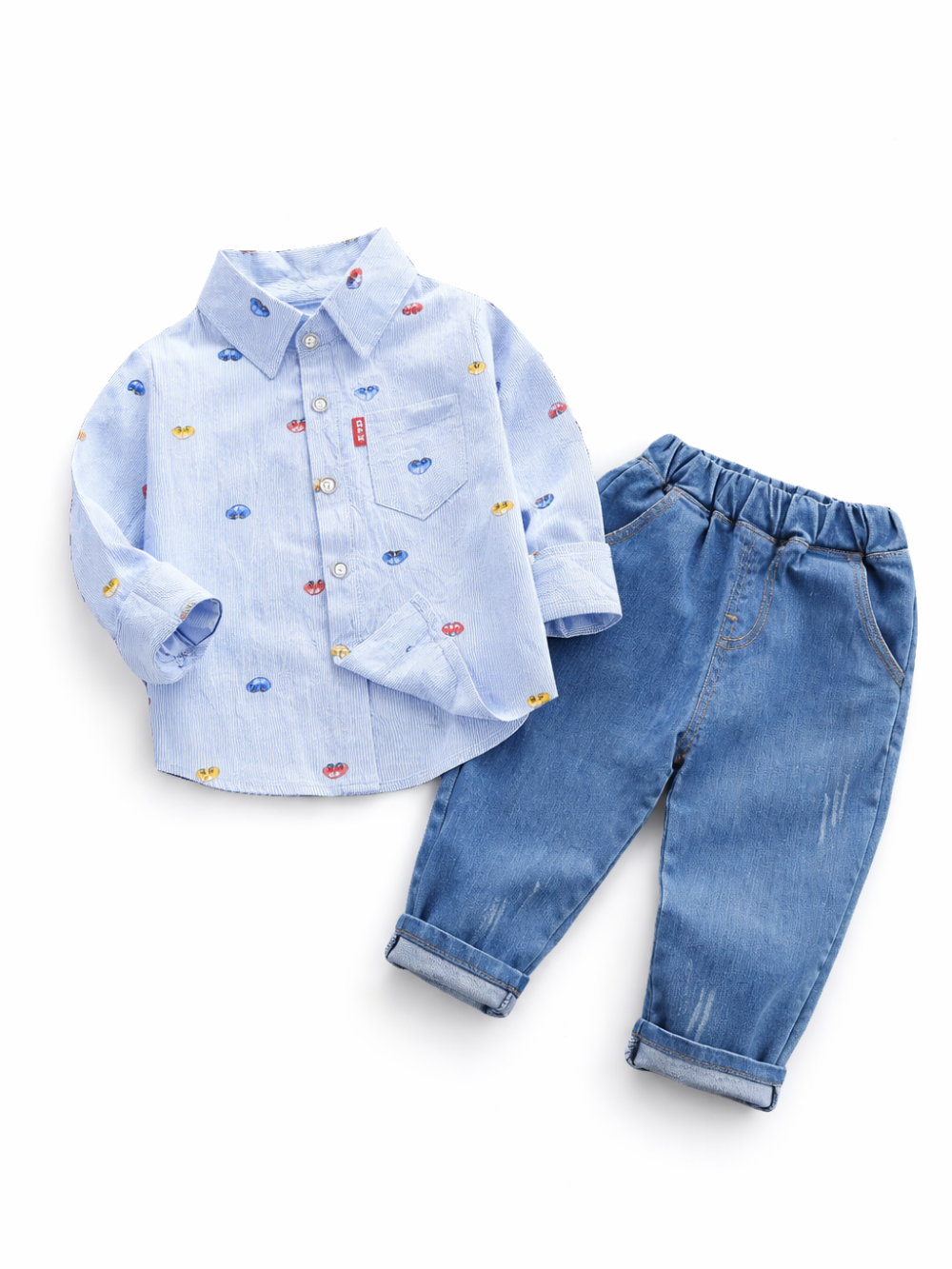 白背景に、子ども用の上下2点セットが平置きで配置されている。上は淡いブルーの長袖ボタンシャツで、小さなカラフルな車の柄が散りばめられ、襟と胸ポケット、白いボタン、赤いタグが付いている。下はミディアムブルーのデニムパンツで、ウエストはゴム仕様、裾はロールアップされている。全体が均一な光で撮影され、素材の質感と輪郭がはっきり見える。
