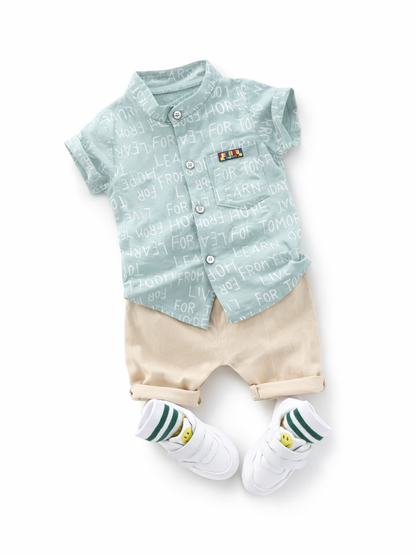 白背景に、子ども用コーディネート一式が平置きで縦に配置されている。上は淡いミントグリーンの半袖ボタンシャツで、白い文字の総柄プリントと胸ポケットの小さなカラーパッチがある。下はベージュのショートパンツで、裾はロールアップ。足元には白い面ファスナーのスニーカーと、グリーンのライン入りソックス（黄色いスマイル柄）が合わせて置かれている。全体が均一な光で撮影され、輪郭と素材感がはっきり見える。
