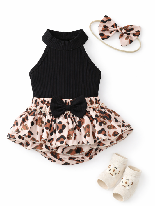 白背景に、ベビー用のコーディネート一式が平置きで配置されている。黒のリブ素材ホルターネックロンパースに、ピンクベージュ系のレオパード柄フリルスカート（中央に黒いリボン付き）が組み合わさっている。右上には同柄のリボンヘアバンド、右下にはオフホワイトのベビーソックス（くまの刺繍入り）が置かれている。全体が均一な光で撮影され、輪郭と素材感がはっきり見える。
