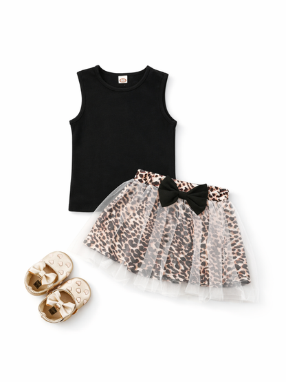 白背景に、子ども用コーディネート一式が平置きで配置されている。上は黒のノースリーブトップスで、丸首のシンプルなデザイン。下はレオパード柄のスカートで、上に白いチュールが重なり、ウエスト部分に黒いリボンが付いている。左下にはゴールド系のリボン付きベビーシューズが並べて置かれている。全体が均一な光で撮影され、輪郭と素材感がはっきり見える。
