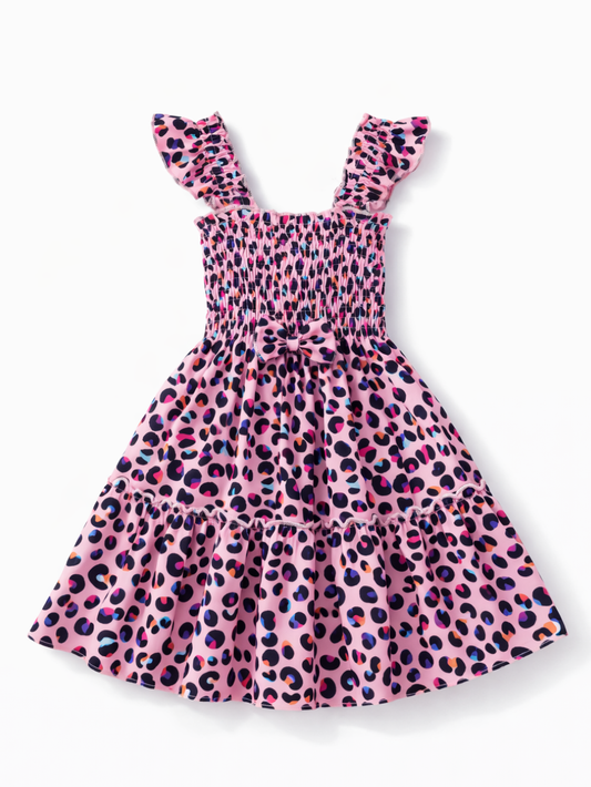 白背景のスタジオで撮影された、ピンクのレオパード柄ティアードワンピース。シャーリング仕様の身頃とフリル付きの肩ストラップ、ウエストの小さなリボンが特徴で、裾は2段のギャザーでふんわり広がっている。
