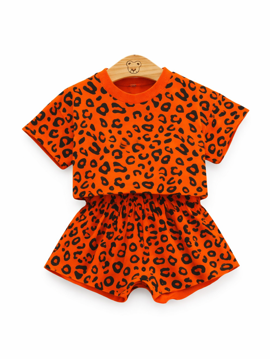 白背景に木製ハンガーで掛けられた、オレンジ色のレオパード柄半袖Tシャツとフリルショートパンツの子ども用上下セット。
