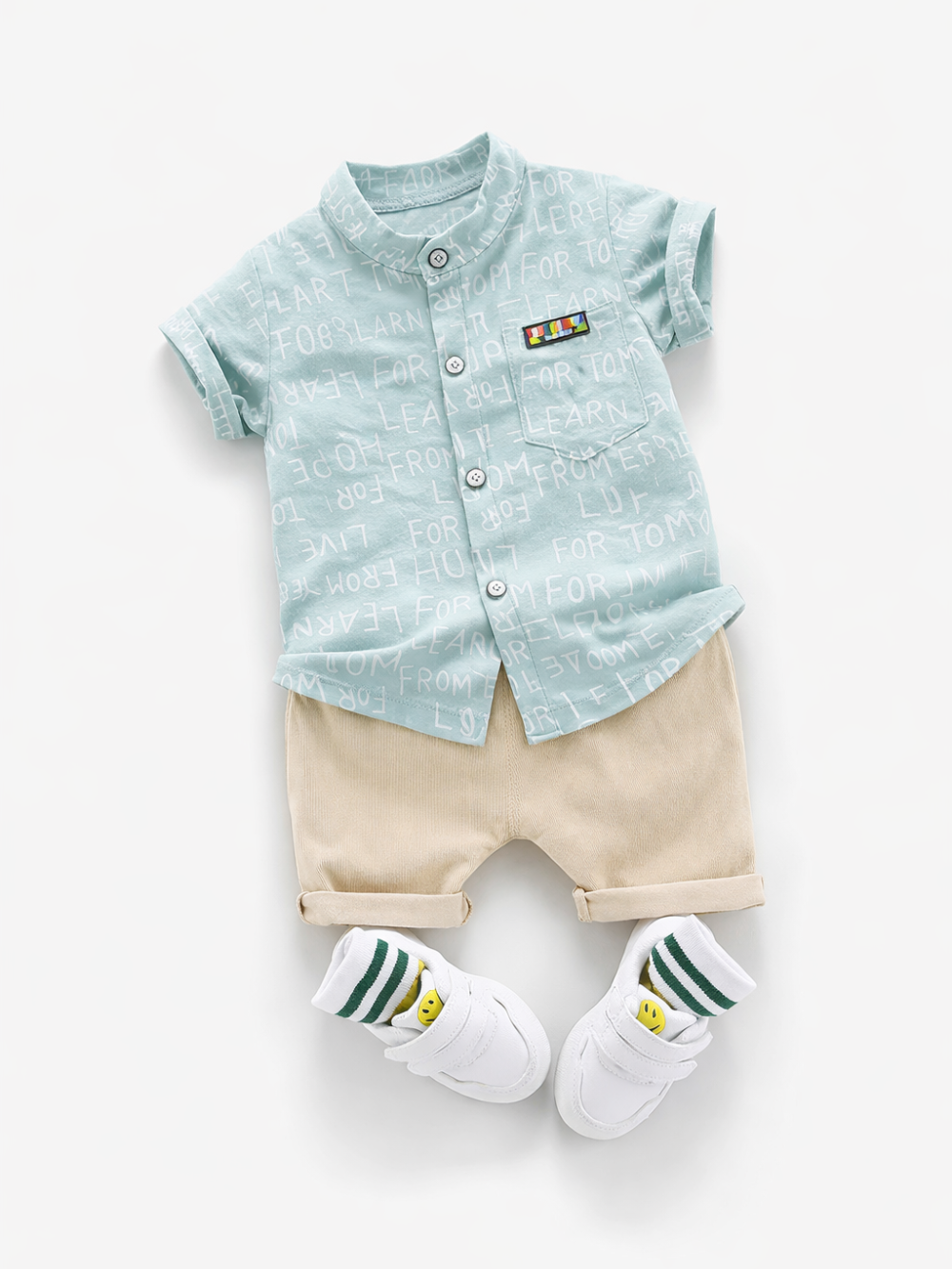 白背景のフラットレイ。淡いミントグリーンの文字柄半袖ボタンシャツ、ベージュのショートパンツ、白いスニーカー、グリーンストライプの靴下（スマイル柄）をセットで配置した幼児用コーデ。
