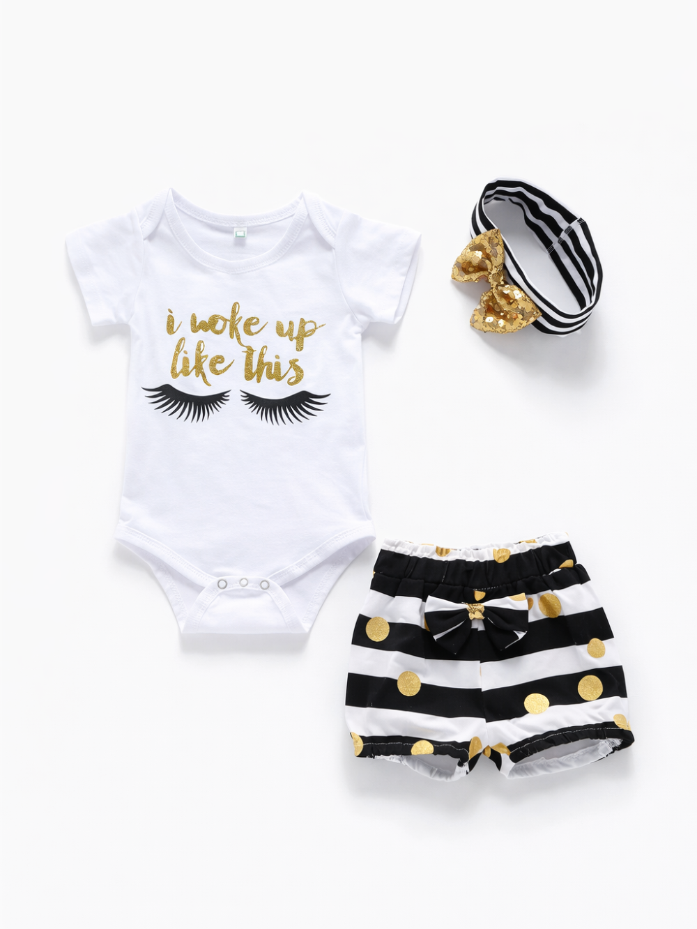 白背景に並べられたベビー用3点セット。金色の「i woke up like this」とまつげプリント入り白いロンパース、黒白ボーダーにゴールドドットのショートパンツ、ゴールドのスパンコールリボン付きボーダー柄ヘアバンド。
