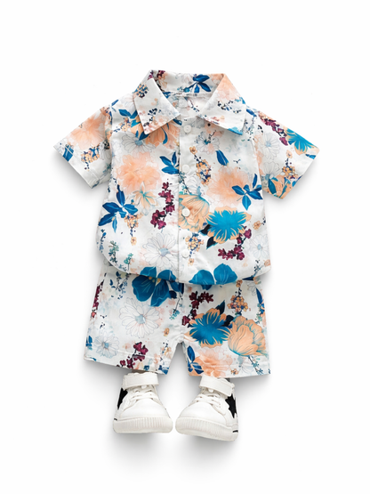 白背景に、白地にブルーとピーチ系の花柄が入った半袖ボタンシャツと同柄ショートパンツのキッズ上下セットが平置きで並び、白いスニーカー（黒い星モチーフ付き）も一緒に写っている。

