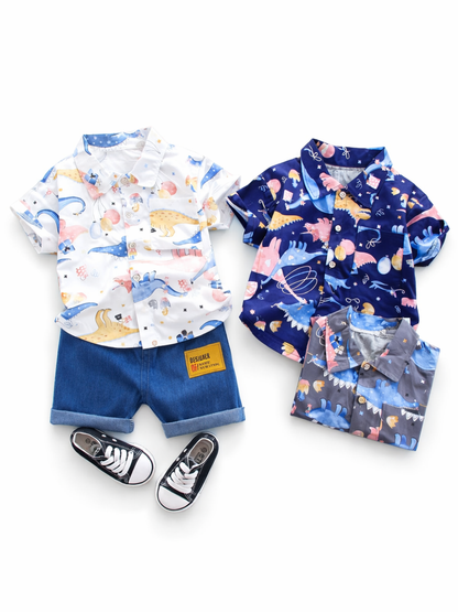 白背景に、恐竜柄のキッズシャツが2点並び、左は白地の恐竜プリント半袖シャツとデニムショートパンツ、黒いスニーカーがセットで写っている。右はネイビー地の恐竜プリント半袖シャツと、下にグレーの同柄シャツがたたんで置かれている。
