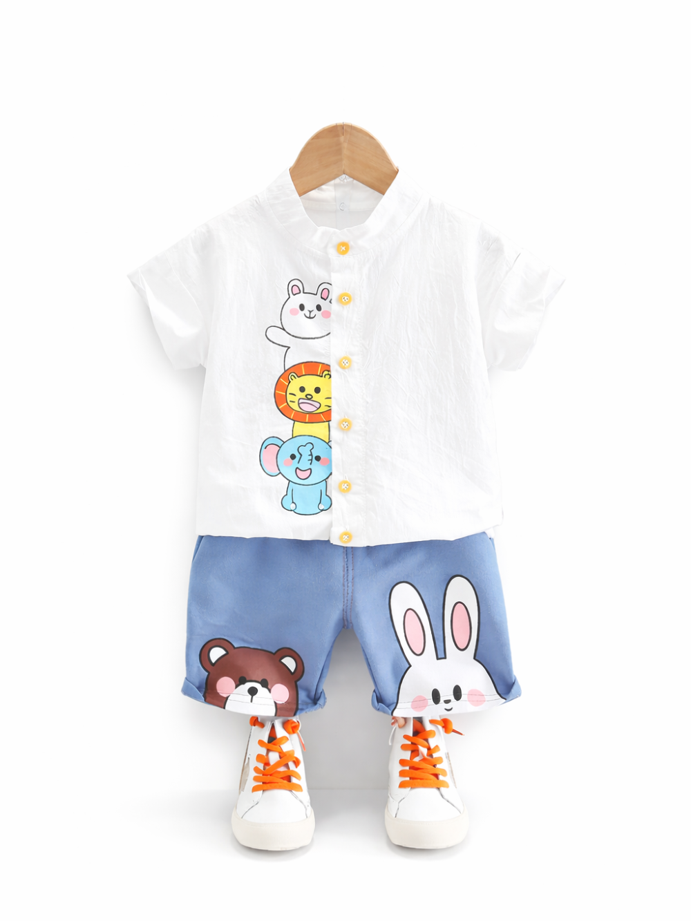 白背景に、木製ハンガーに掛かったキッズの上下セットが写っている。白い半袖ボタンシャツに動物のイラストが入り、ライトブルーのショートパンツにはクマとウサギの顔がプリントされている。足元にはオレンジの靴ひも付きの白いスニーカーが並んでいる。

