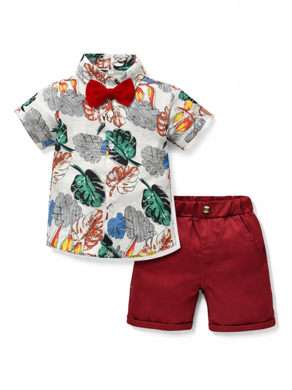 白背景に、キッズの上下セットが写っている。白地にトロピカルリーフ柄の半袖シャツには赤い蝶ネクタイが付いており、右側にボルドーのショートパンツが並んでいる。
