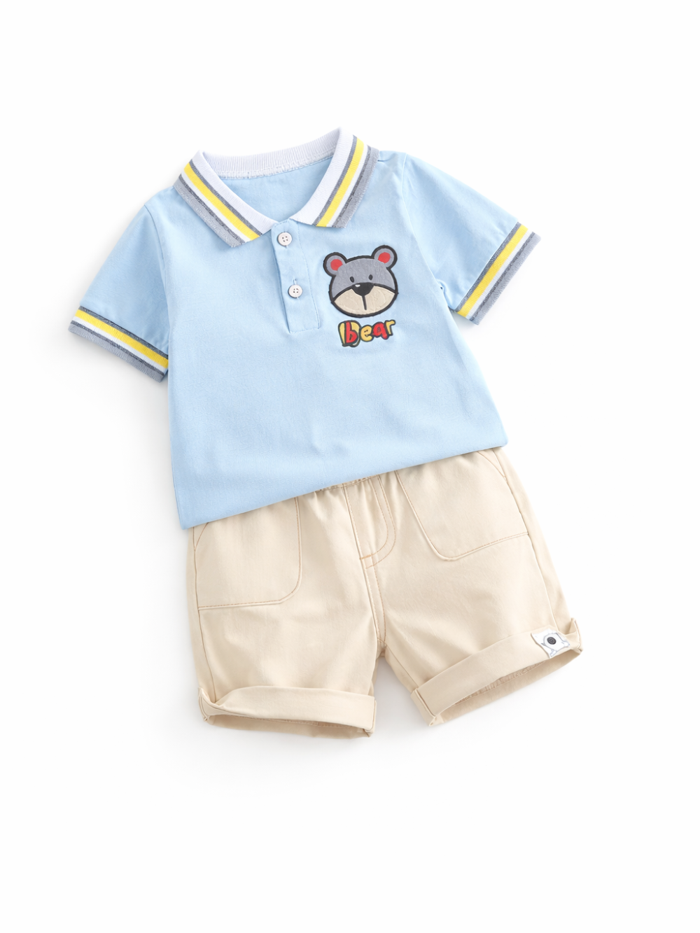 白背景にフラットレイで整えたキッズ用上下2点セット。水色の半袖ポロシャツ（襟・袖口にライン、胸にくまワッペン）と、ベージュのショートパンツ（ロールアップ裾・ポケット付き）をカタログ風に配置している。
