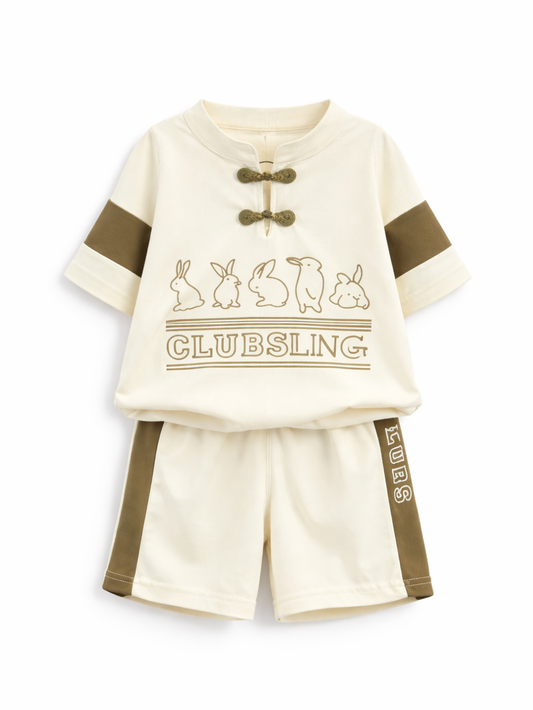 白背景に並べたキッズ用上下2点セット。アイボリーの半袖トップスはチャイナ風のトグル留め付きで、うさぎのイラストと「CLUBSLING」ロゴ入り。合わせてアイボリーのショートパンツにカーキのサイドパネルと文字プリントがある。
