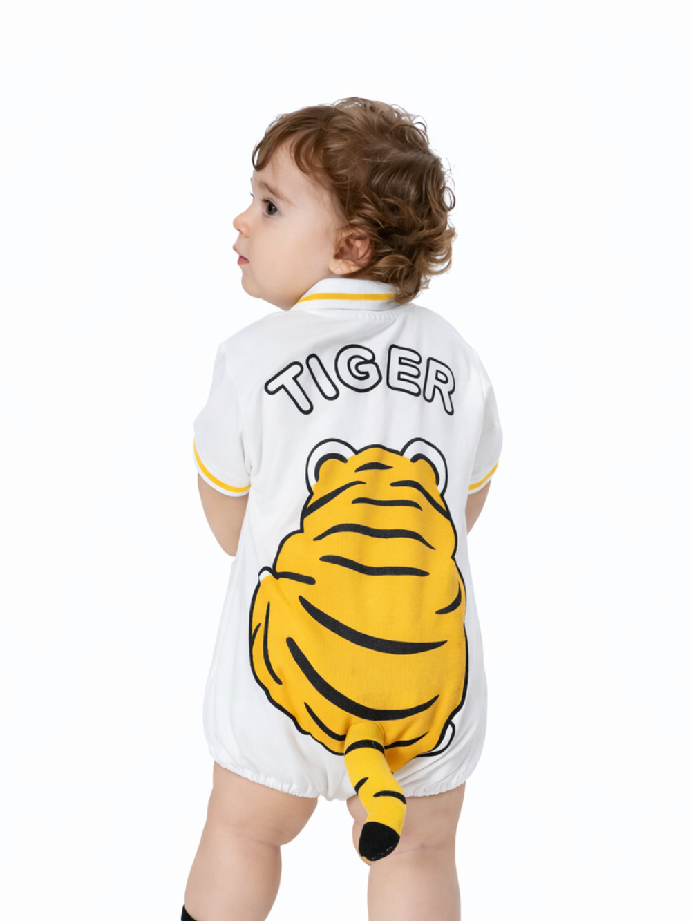 白背景で後ろ向きに立つ幼児。白いロンパースの背中に「TIGER」の文字と大きなトラのイラスト、立体のしっぽが付いており、黄色のライン入りの襟と袖が特徴。振り返るポーズで撮影されている。
