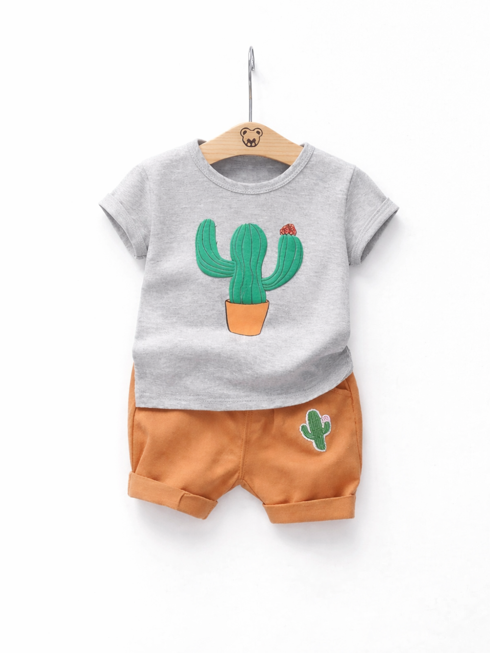 白背景に木製ハンガーで掛けたキッズ半袖セットアップ。グレーのサボテンプリントTシャツと、サボテンワッペン付きオレンジのショートパンツ。
