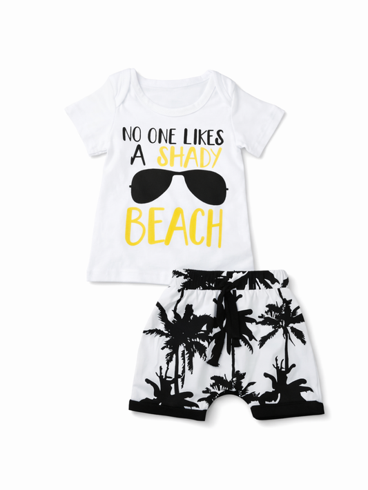 白背景に配置されたキッズ用夏服2点セット。白い半袖Tシャツにサングラスのイラストと英字プリント、黒×白のヤシの木柄ショートパンツ（ウエストゴム、紐付き）がセットになっている。

