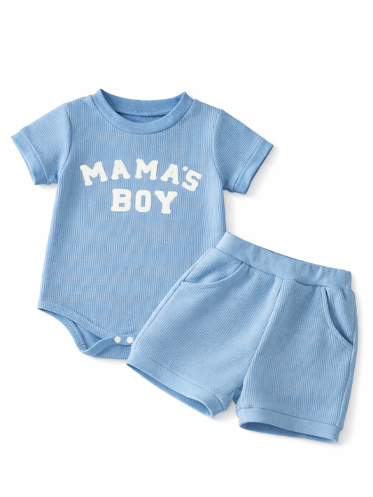 白背景に平置きされたベビー男の子用の上下セット。ライトブルーのリブ素材ロンパースに「MAMA’S BOY」の白文字プリント、同素材のショートパンツがセットになっている。やわらかな陰影で立体感が出ている。
