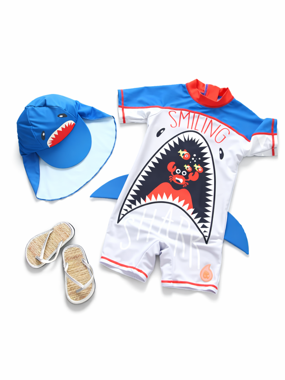 白背景に平置きされたキッズ用シャーク柄スイムウェアセット。赤い襟と青い袖の半袖ワンピース水着に、サメの口デザインと「SMILING」文字、カニのイラストが入っている。左にサメ顔のつば広ハット、下にビーチサンダルが配置されている。
